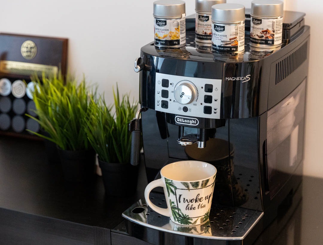 Czarny ekspres do kawy DeLonghi Magnifica S z filiżanką z napisem 'I woke up like this' i czterema puszkami kawy smakowej Deluxe na górze, obok doniczki ze sztuczną trawą.