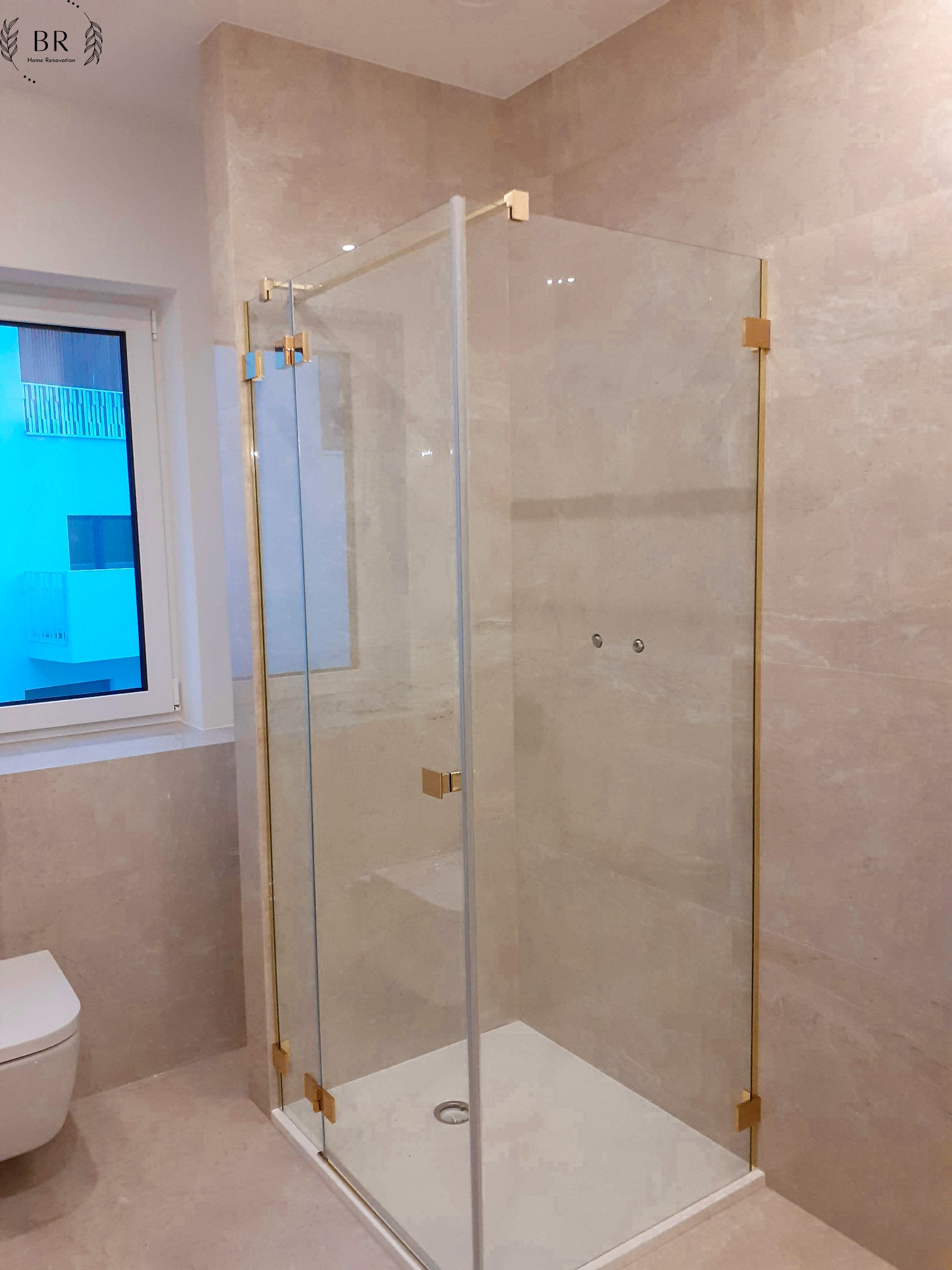 Nowoczesna kabina prysznicowa z przezroczystego szkła z złotymi okuciami, zamontowana w łazience wykończonej beżowymi płytkami imitującymi marmur, widoczny fragment toalety i okno z odbiciem...