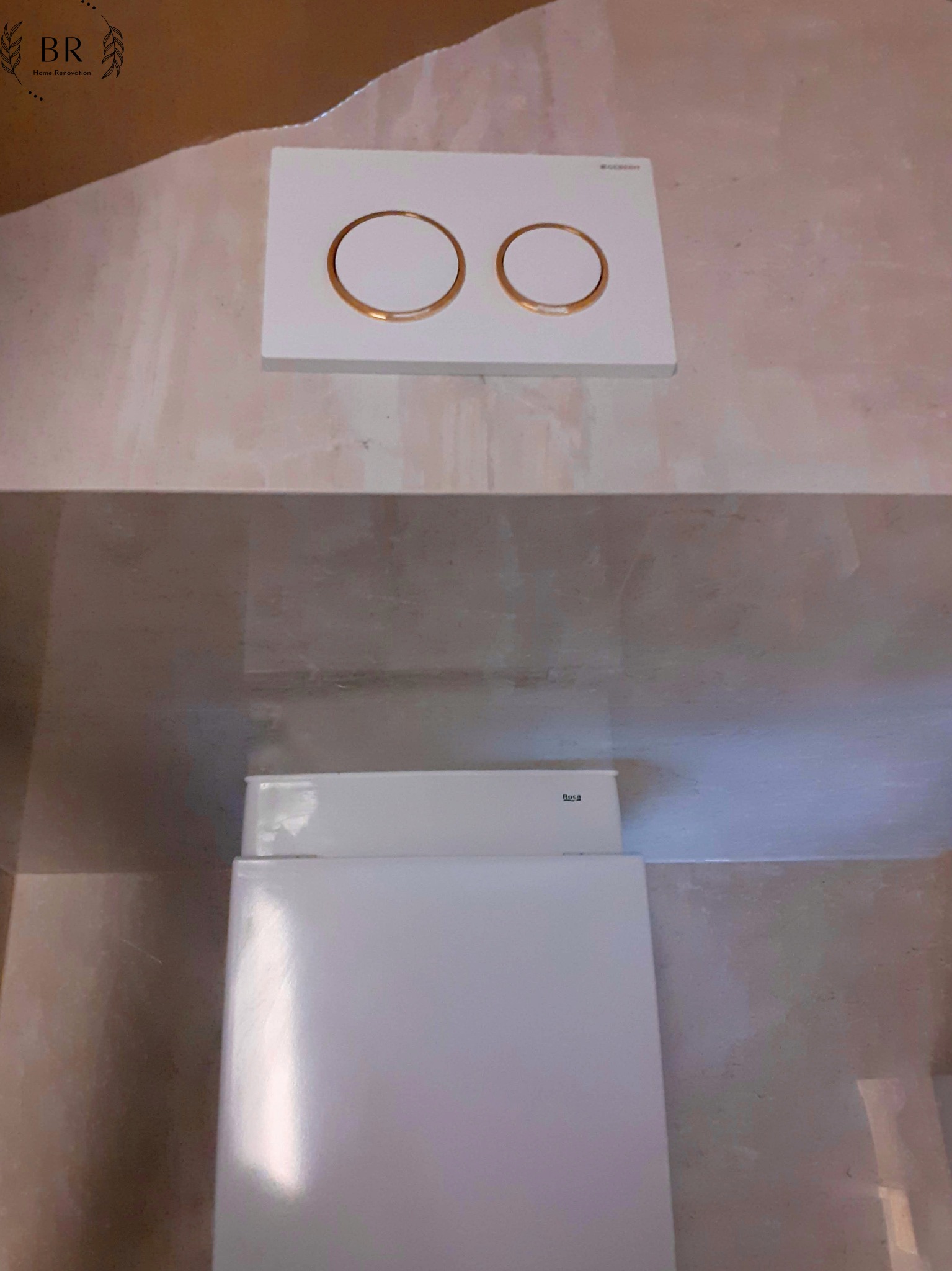 Wisząca toaleta z białym panelem spłukującym z dwoma złotymi okręgami na tle beżowych płytek ściennych.