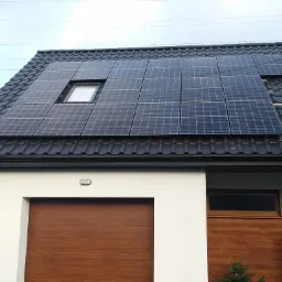 Panele słoneczne na dachu domu jednorodzinnego z czarną dachówką, widoczna drabina oparta o dach, element instalacji na kominie, okno dachowe.