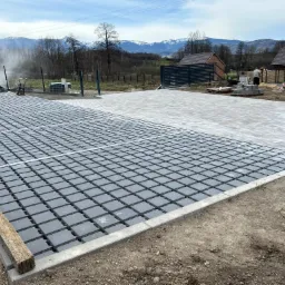 Częściowo ułożona nawierzchnia z ażurowych płyt betonowych na podsypce, widoczni pracownicy i sprzęt budowlany, w tle zabudowa jednorodzinna i zalesione wzgórza.