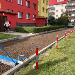 Przygotowanie podłoża pod kostkę brukową przy bloku mieszkalnym: wyrównywanie terenu specjalistycznym narzędziem, widoczne obrzeża i palety z kostką, w tle budynki w czerwonej i żółtej kolorystyce.