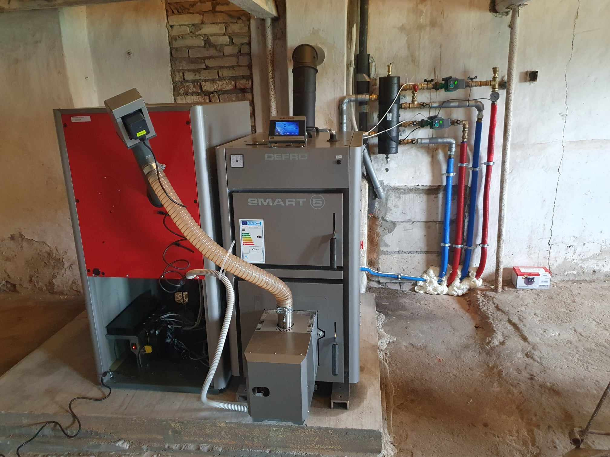 Nowoczesny piec Defro Smart E z podajnikiem, widoczne rury instalacji grzewczej w kolorach niebieskim i czerwonym, oraz regulator temperatury na górze pieca.