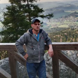 Mężczyzna w szarej bluzie z kapturem i dżinsach opiera się o drewnianą barierkę na tle rozległego krajobrazu zalesionych wzgórz i oddalonej wioski.