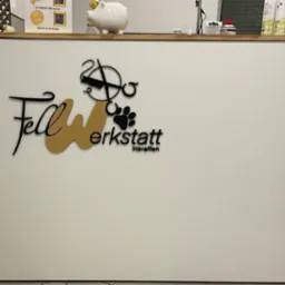 Białe biurko z czarnym i złotym logo 'Fellwerkstatt Horaffen' z motywami zwierzęcymi i fryzjerskimi. Nad biurkiem skarbonka świnka i produkty.