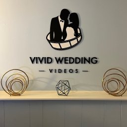 Fragola Creative Studio - Ściana z logo Vivid Wedding Videos, przedstawiającym parę w taśmie filmowej. Dekoracje na półce dodają elegancji. Minimalistyczny design w czerni i złocie.