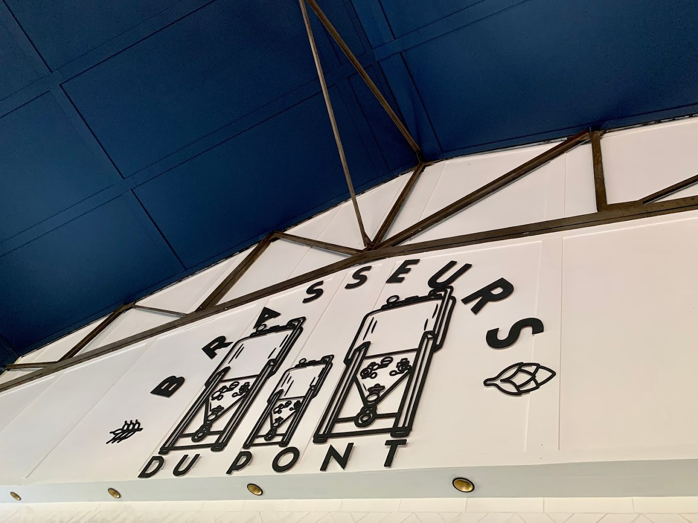 Czarny, przestrzenny napis 'BRASSEURS DUPONT' z graficznymi elementami na białej ścianie pod ciemnoniebieskim sufitem z metalową konstrukcją.