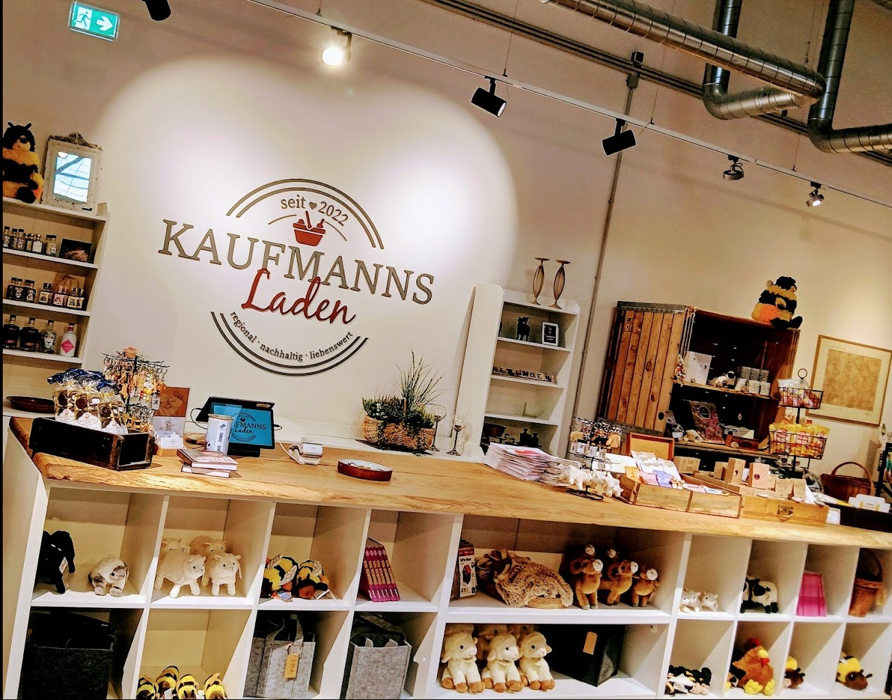 Wnętrze sklepu Kaufmanns Laden z logo na ścianie, drewnianym blatem i półkami wypełnionymi produktami regionalnymi i rękodziełem. Widoczne oświetlenie i elementy dekoracyjne.