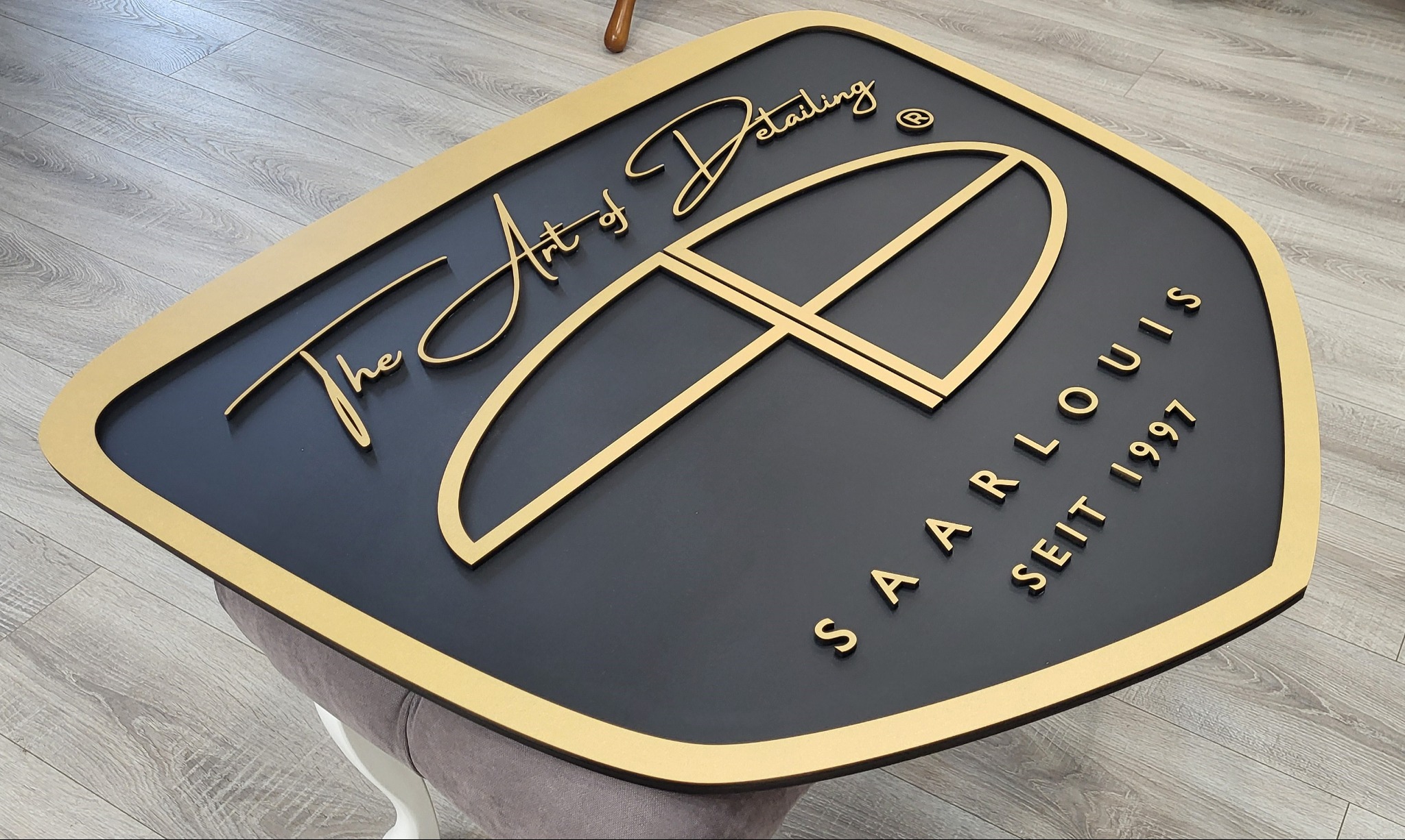 Elegancki szyld firmowy z logo 'The Art of Detailing Saarlouis seit 1997' w kolorze złotym na czarnym tle, umieszczony na szarym siedzisku, widok z góry.