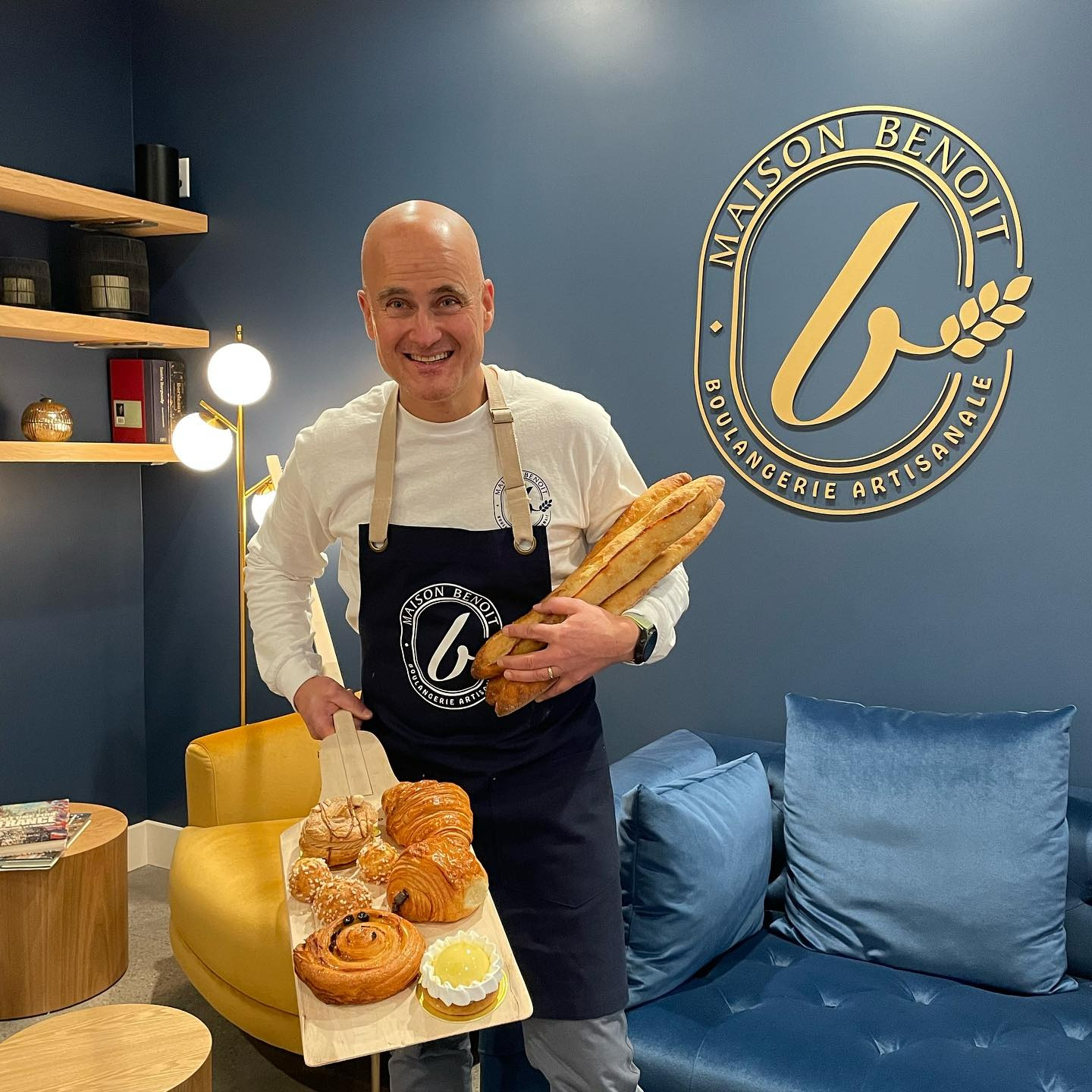 Uśmiechnięty piekarz w fartuchu prezentuje pieczywo na desce i bagietki, na tle ściany z logo 'Maison Benoit Boulangerie Artisanale'.