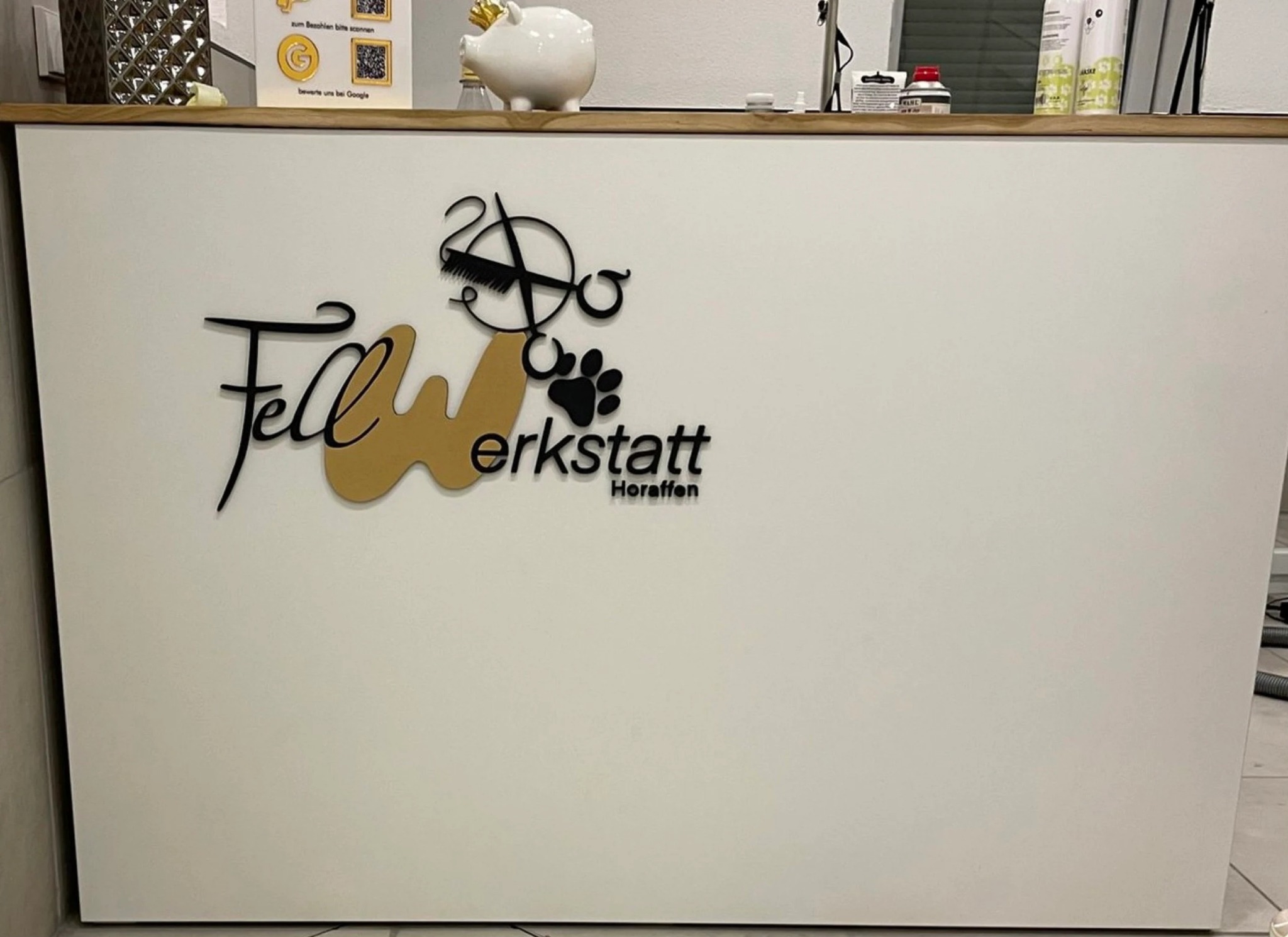 Białe biurko z czarnym i złotym logo 'Fellwerkstatt Horaffen' z motywami zwierzęcymi i fryzjerskimi. Nad biurkiem skarbonka świnka i produkty.