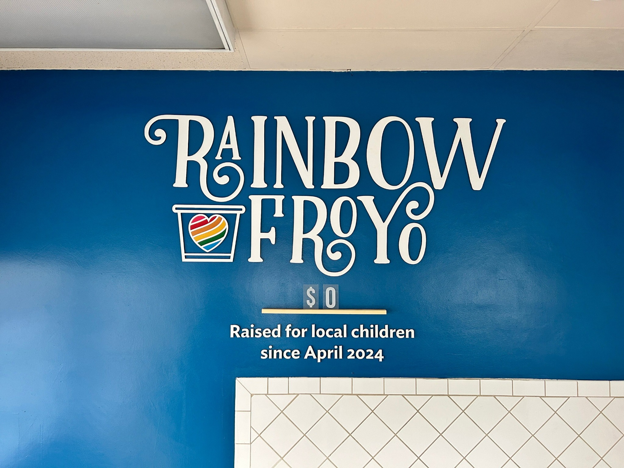 Napis 'Rainbow FroYo' na niebieskiej ścianie z logo w kształcie tęczowego serca w kubku. Poniżej informacja o zbiórce pieniędzy dla lokalnych dzieci od kwietnia 2024.