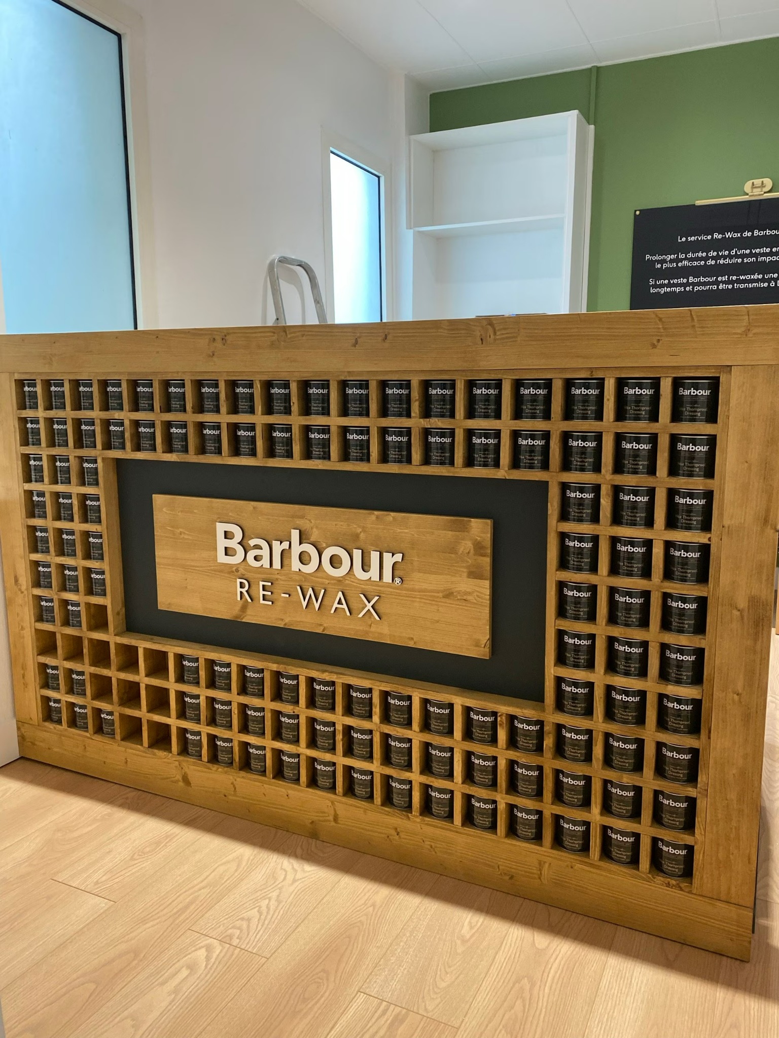 Ekspozycja produktów Barbour Re-Wax na drewnianym regale z logo, w jasnym wnętrzu sklepu. Prezentacja marki i usługi odnawiania odzieży woskowanej.