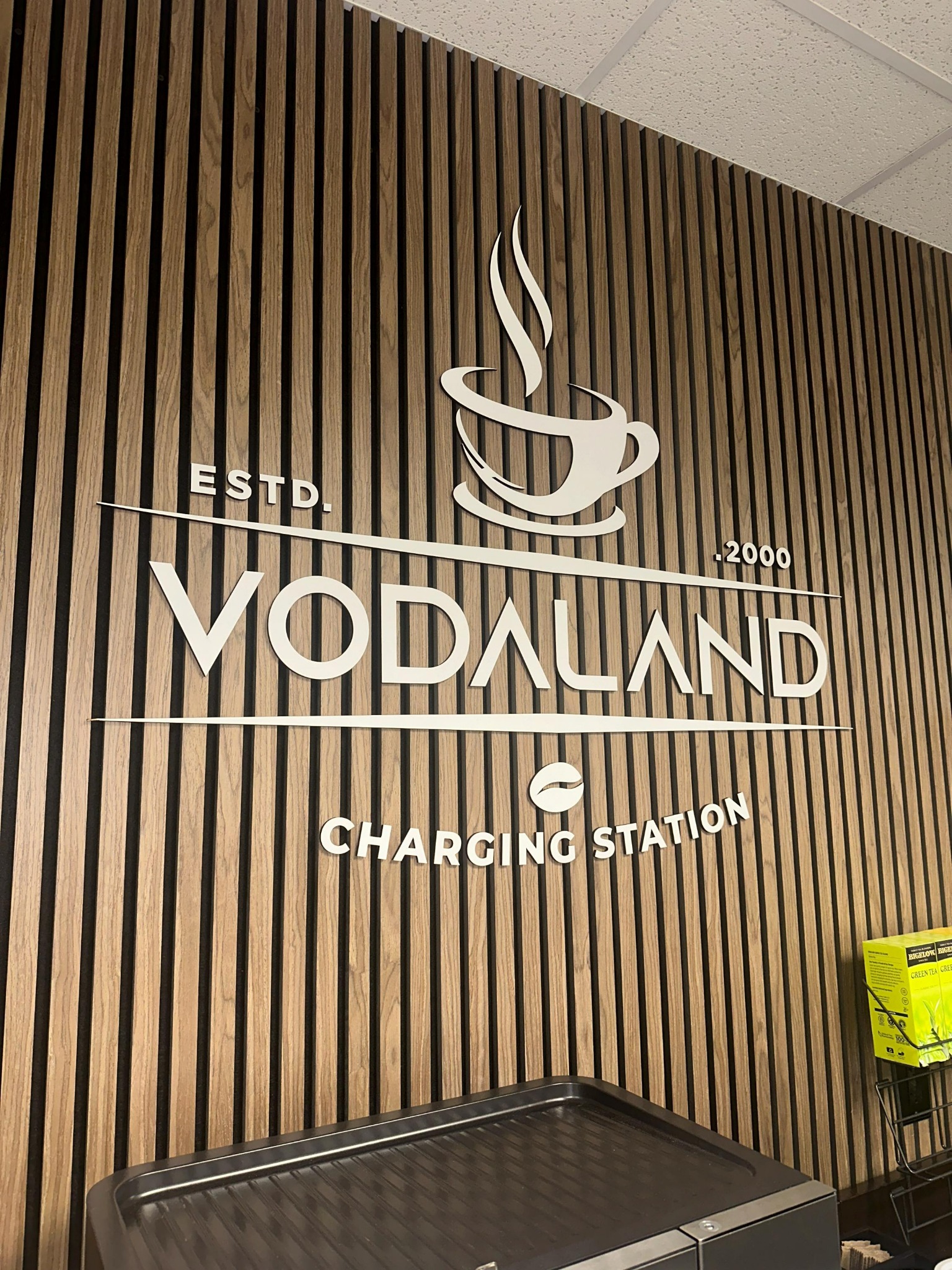 Ściana z drewnianych paneli z białym logo 'VODALAND Charging Station' z motywem filiżanki kawy. Nowoczesny design, ciepłe oświetlenie, widoczny ekspres do kawy.