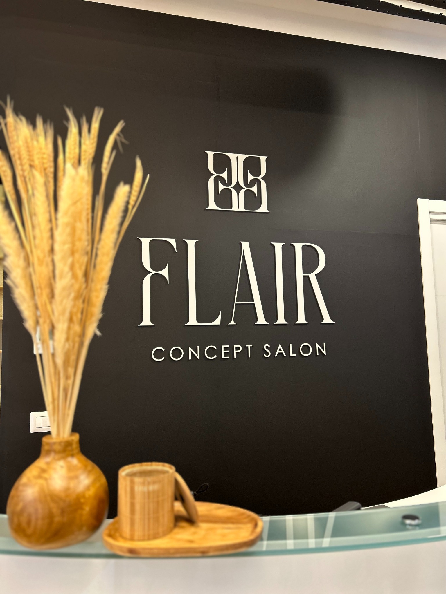 Logo 'Flair Concept Salon' na czarnej ścianie, z dekoracjami z drewna i suszonych traw na pierwszym planie. Minimalistyczny design, ciepłe oświetlenie.