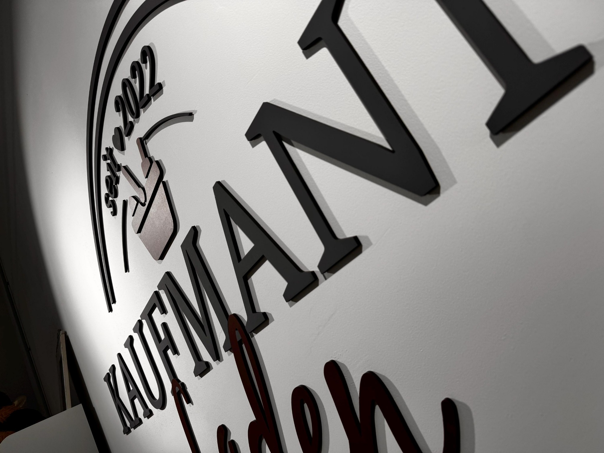 Czarno-brązowy, trójwymiarowy napis 'Kaufman' z motywem roślinnym i rokiem '2022', umieszczony na białej ścianie, widziany pod kątem.