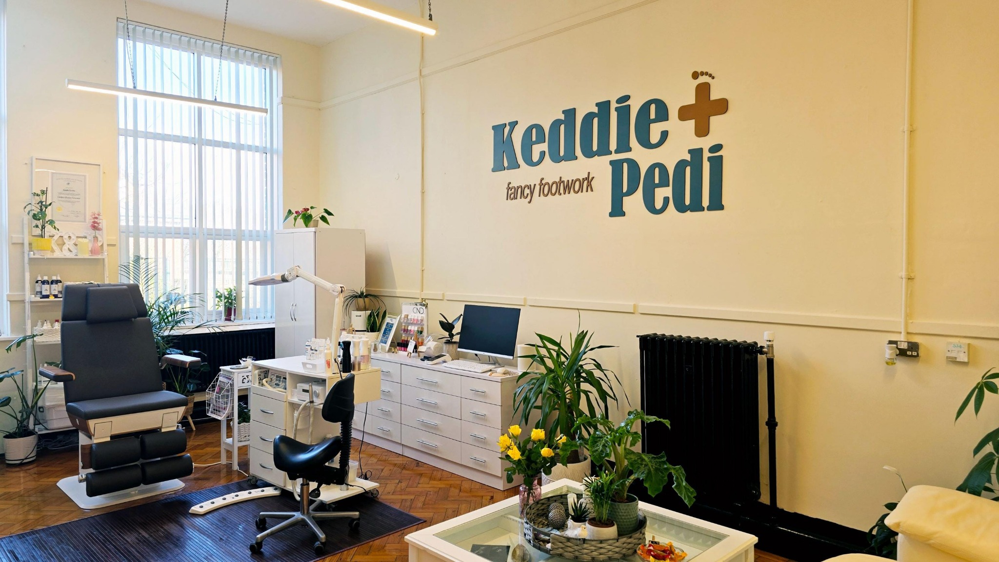 Wnętrze gabinetu podologicznego: fotel zabiegowy, biurko z komputerem, rośliny doniczkowe i logo firmy na ścianie. Jasne, przestronne pomieszczenie z parkietem.