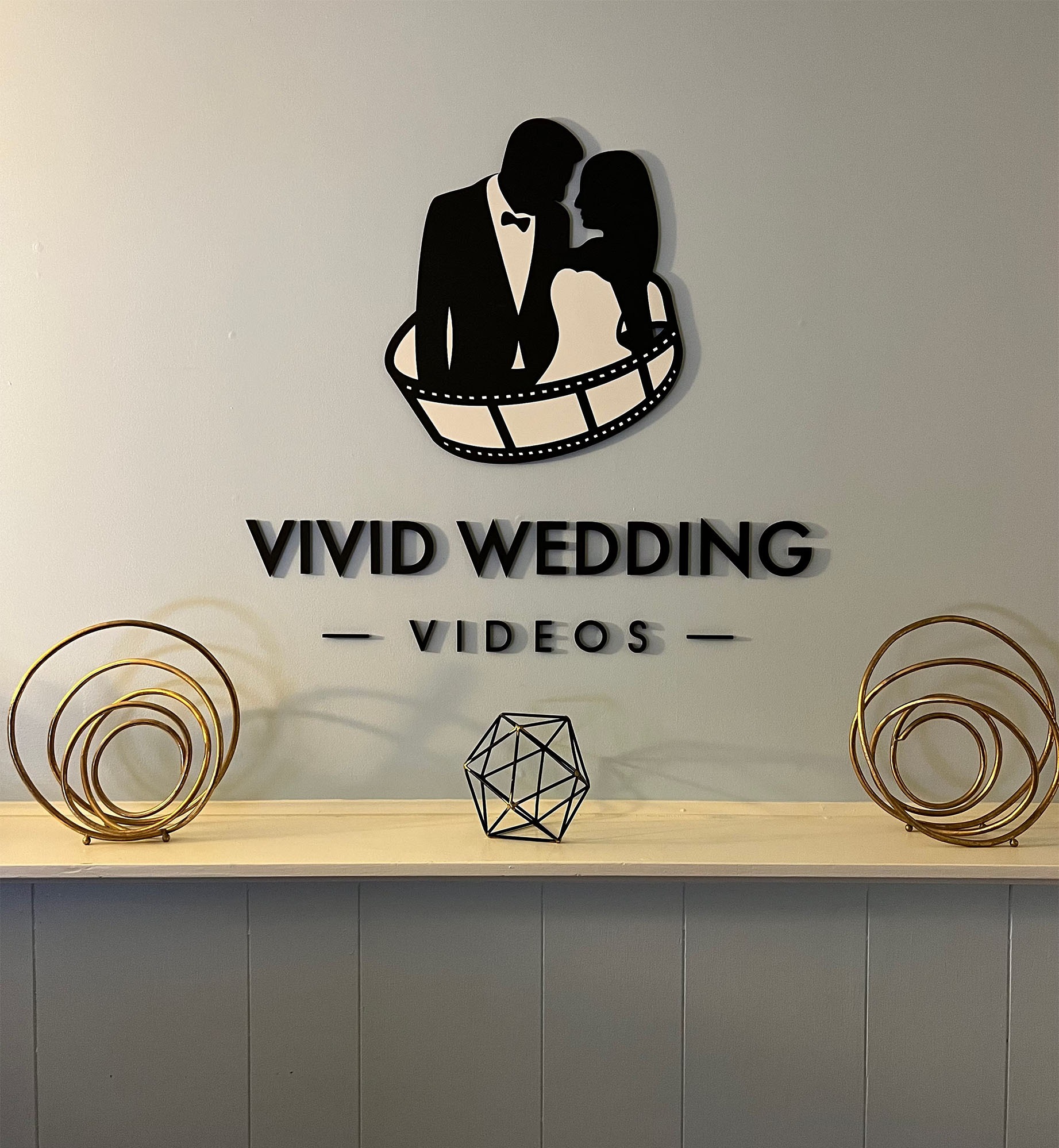 Ściana z logo Vivid Wedding Videos, przedstawiającym parę w taśmie filmowej. Dekoracje na półce dodają elegancji. Minimalistyczny design w czerni i złocie.