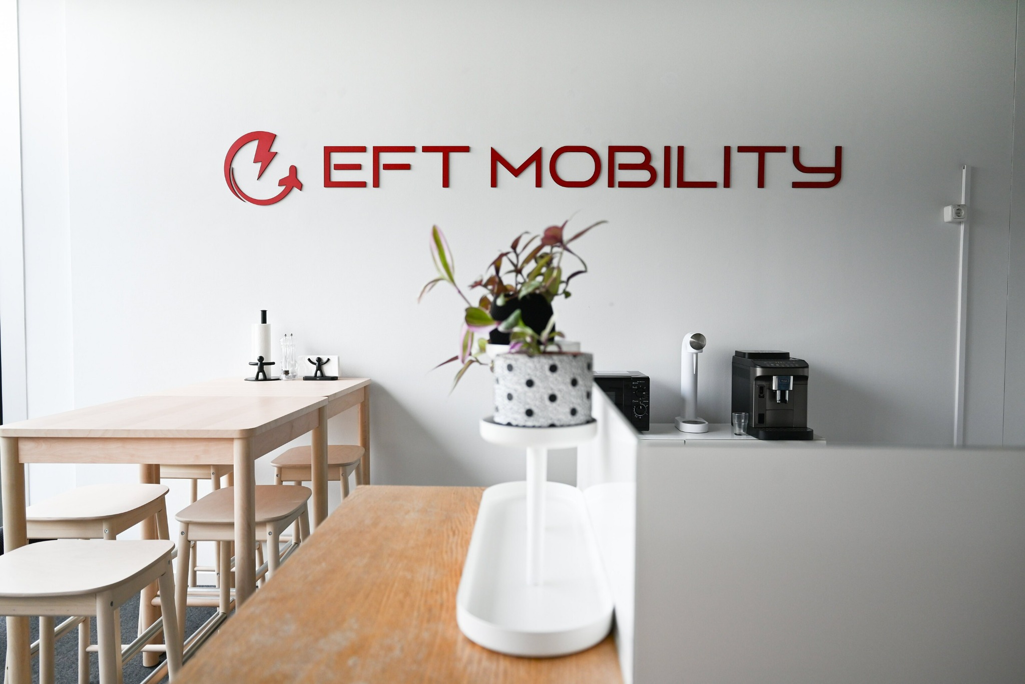 Nowoczesne biuro z czerwonym logo 'EFT Mobility' na białej ścianie. Jasne drewniane meble, minimalistyczny wystrój. Roślina doniczkowa dodaje świeżości.
