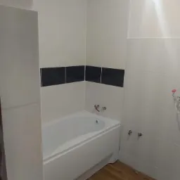 Wyremontowana łazienka z białą wanną, jasnymi płytkami i poziomym pasem czarnych płytek dekoracyjnych oraz drewnopodobną podłogą.