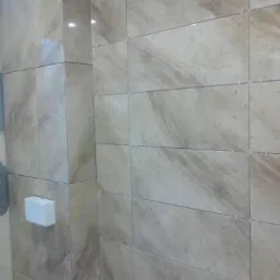 Ściana w trakcie wykańczania z beżowych, marmurkowych płytek ceramicznych ułożonych poziomo, z widocznymi krzyżykami dystansowymi i puszką elektryczną.