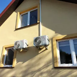 Dwa klimatyzatory marki Rotenso zamontowane na elewacji domu o żółtej barwie, widoczne okna z roletami i odbicia drzew w szybach.