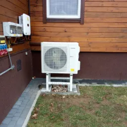 Biała jednostka zewnętrzna pompy ciepła Innova zamontowana na metalowym stelażu przy brązowej drewnianej ścianie domu. Widoczne przyłącza elektryczne i skrzynki na elewacji. Pod stelażem wysypane...