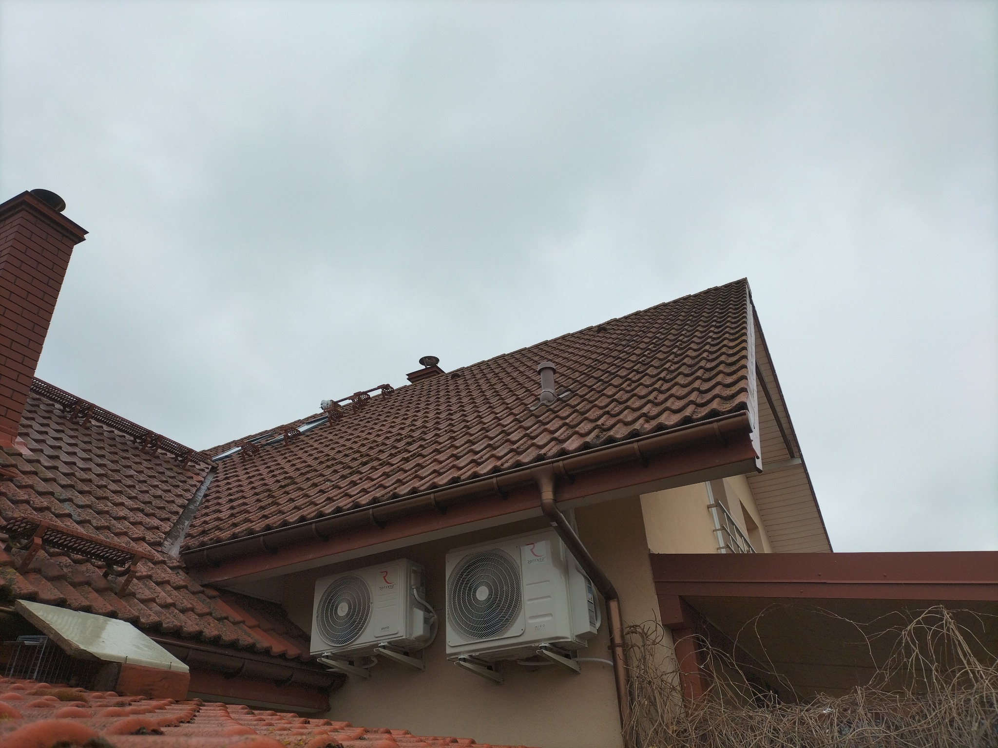 Dwa białe klimatyzatory zamontowane na elewacji domu z brązową dachówką, widoczne rynny i komin na tle pochmurnego nieba.