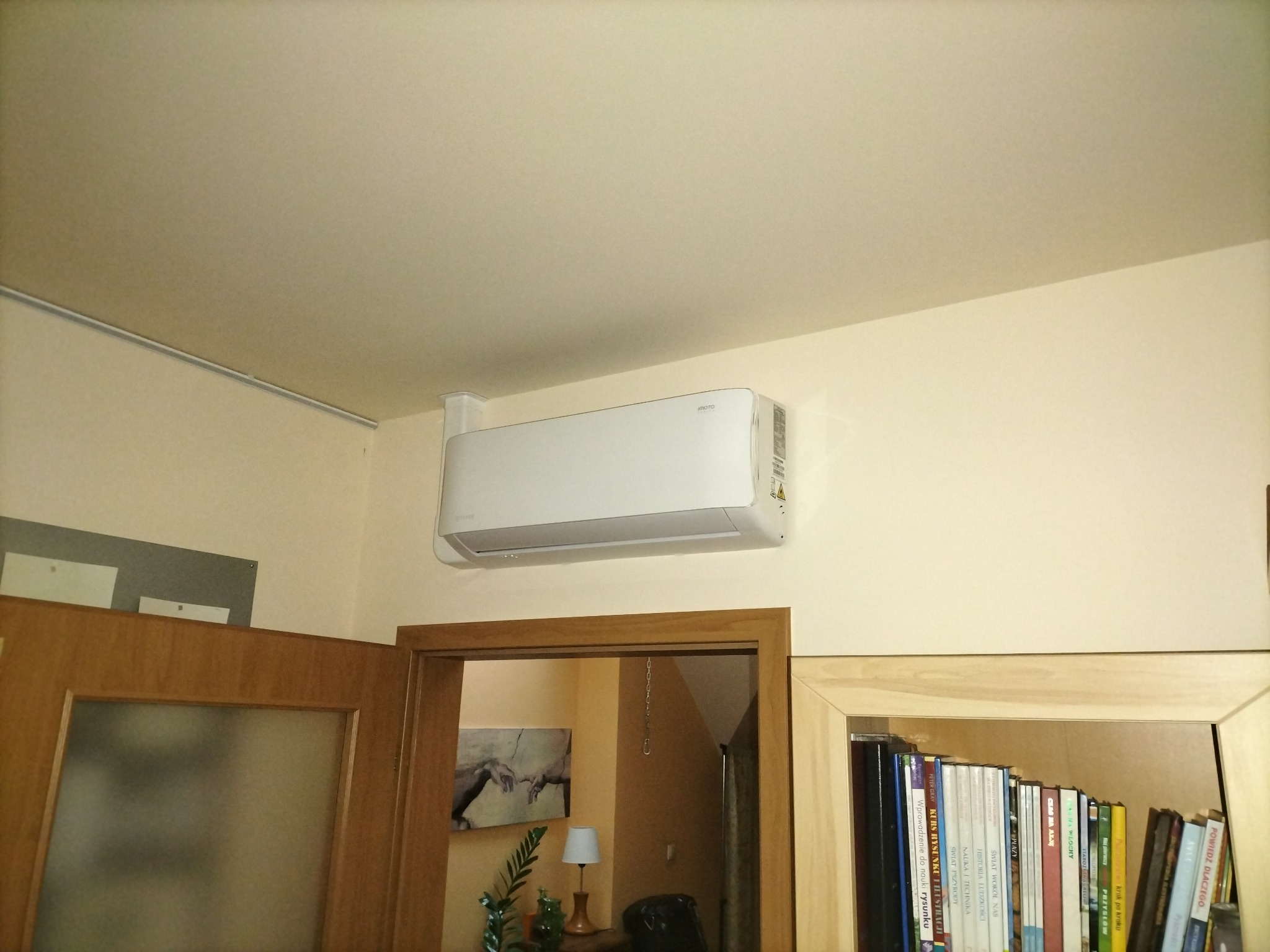 Biała jednostka klimatyzacyjna zamontowana na ścianie w pokoju z widocznymi drzwiami i regałem z książkami. W tle widoczny obraz na ścianie.