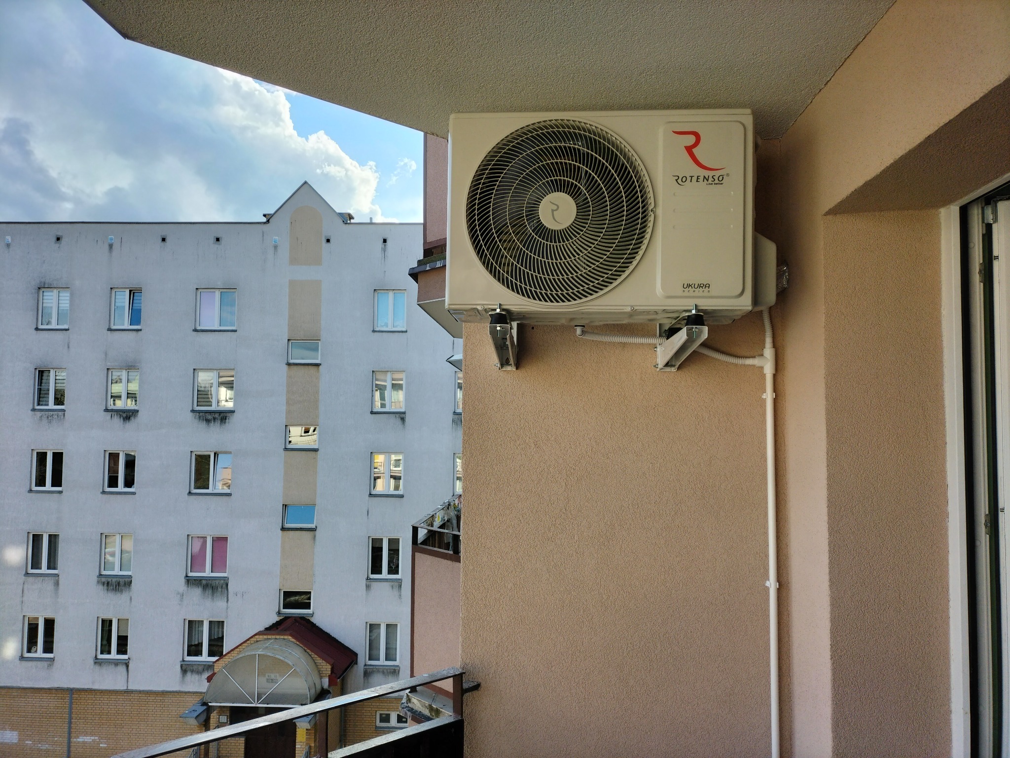 Zewnętrzna jednostka klimatyzacji Rotenso zamontowana na balkonie bloku mieszkalnego, widok na budynek z oknami i błękitne niebo z chmurami.