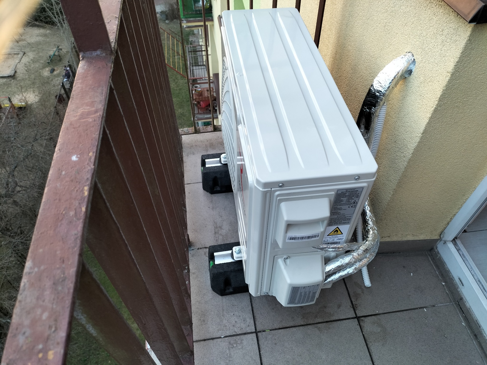 Jednostka zewnętrzna klimatyzatora zamontowana na balkonie, widoczne przewody w izolacji aluminiowej, wsporniki antywibracyjne i metalowa balustrada.