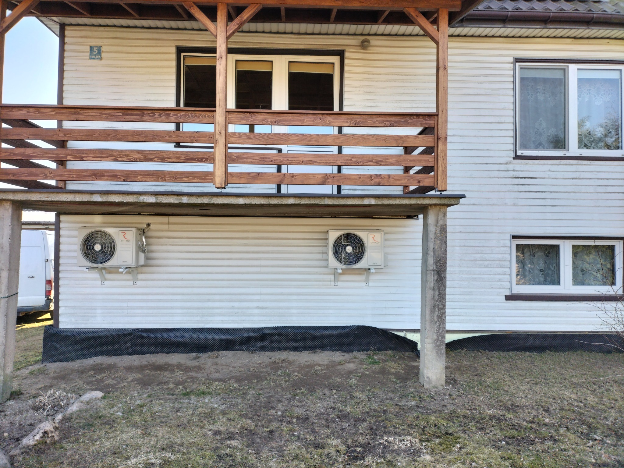 Elewacja domu z dwoma zamontowanymi klimatyzatorami na parterze, widoczny drewniany balkon i część ogrodu.