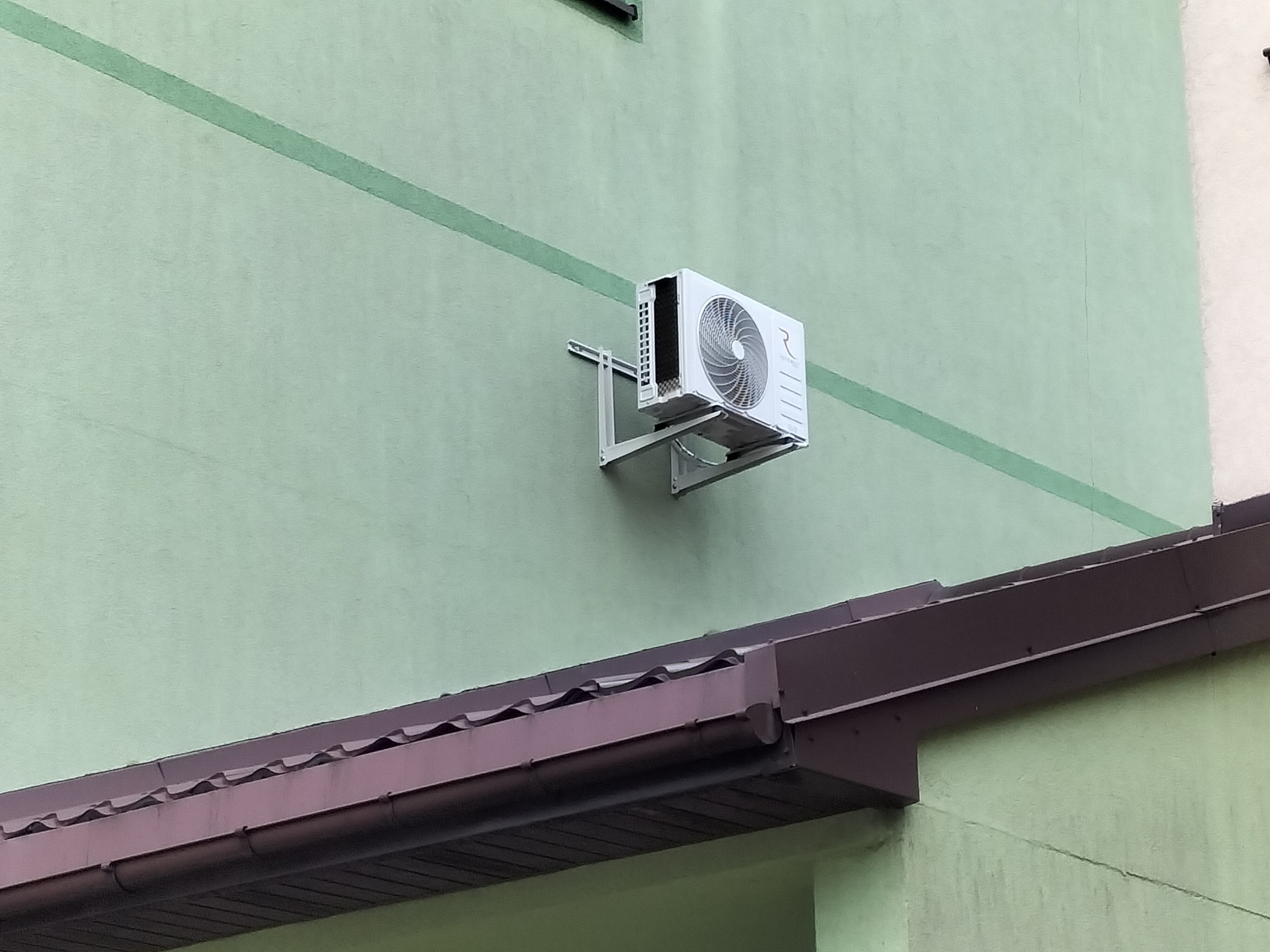 Biała jednostka zewnętrzna klimatyzatora Rotenso Elis zamontowana na jasnym, zielonym budynku z widoczną brązową rynną.