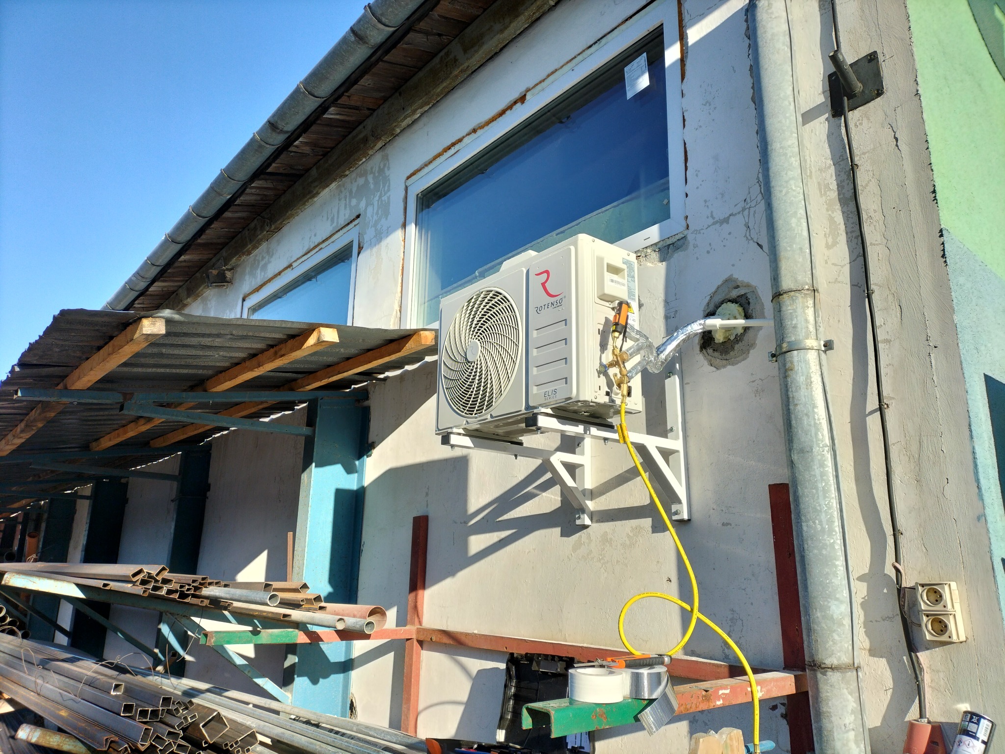 Zewnętrzna jednostka klimatyzatora Rotenso Elis zamontowana na ścianie budynku, widoczne przewody i elementy instalacji, pod spodem rusztowanie z materiałami budowlanymi.
