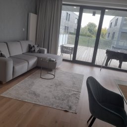 Przybysz - Jasny salon z narożną kanapą, stolikiem kawowym na okrągłych nóżkach i wzorzystym dywanem, widok na taras z meblami ogrodowymi przez duże okno balkonowe.