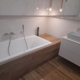 Przybysz - Nowoczesna łazienka z białą wanną obudowaną drewnem, drewnianą podłogą i białą umywalką na szafce. Nad wanną bateria wannowa ze słuchawką prysznicową. Na blacie obok wanny stoi czarna butelka...