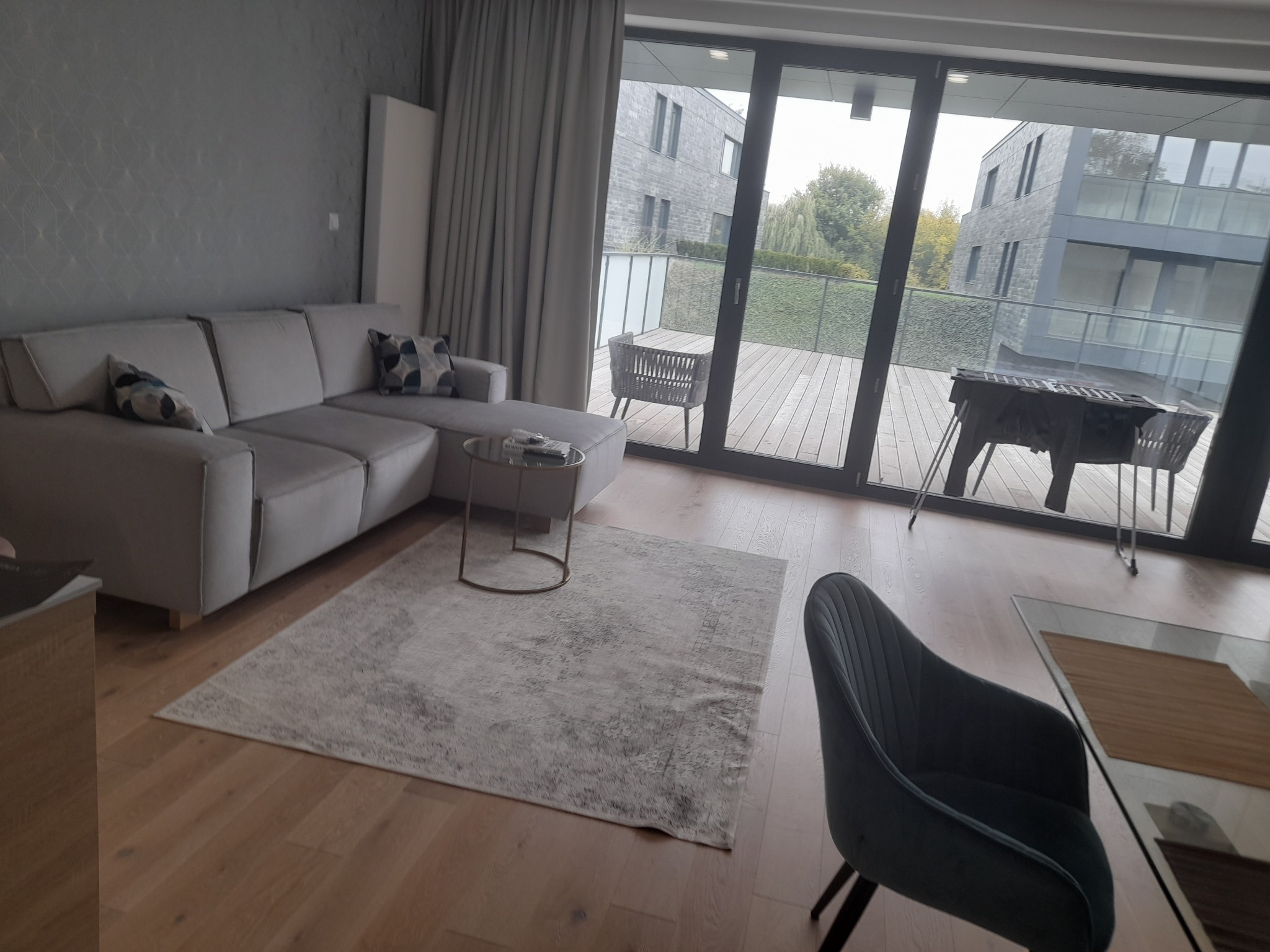 Jasny salon z narożną kanapą, stolikiem kawowym na okrągłych nóżkach i wzorzystym dywanem, widok na taras z meblami ogrodowymi przez duże okno balkonowe.