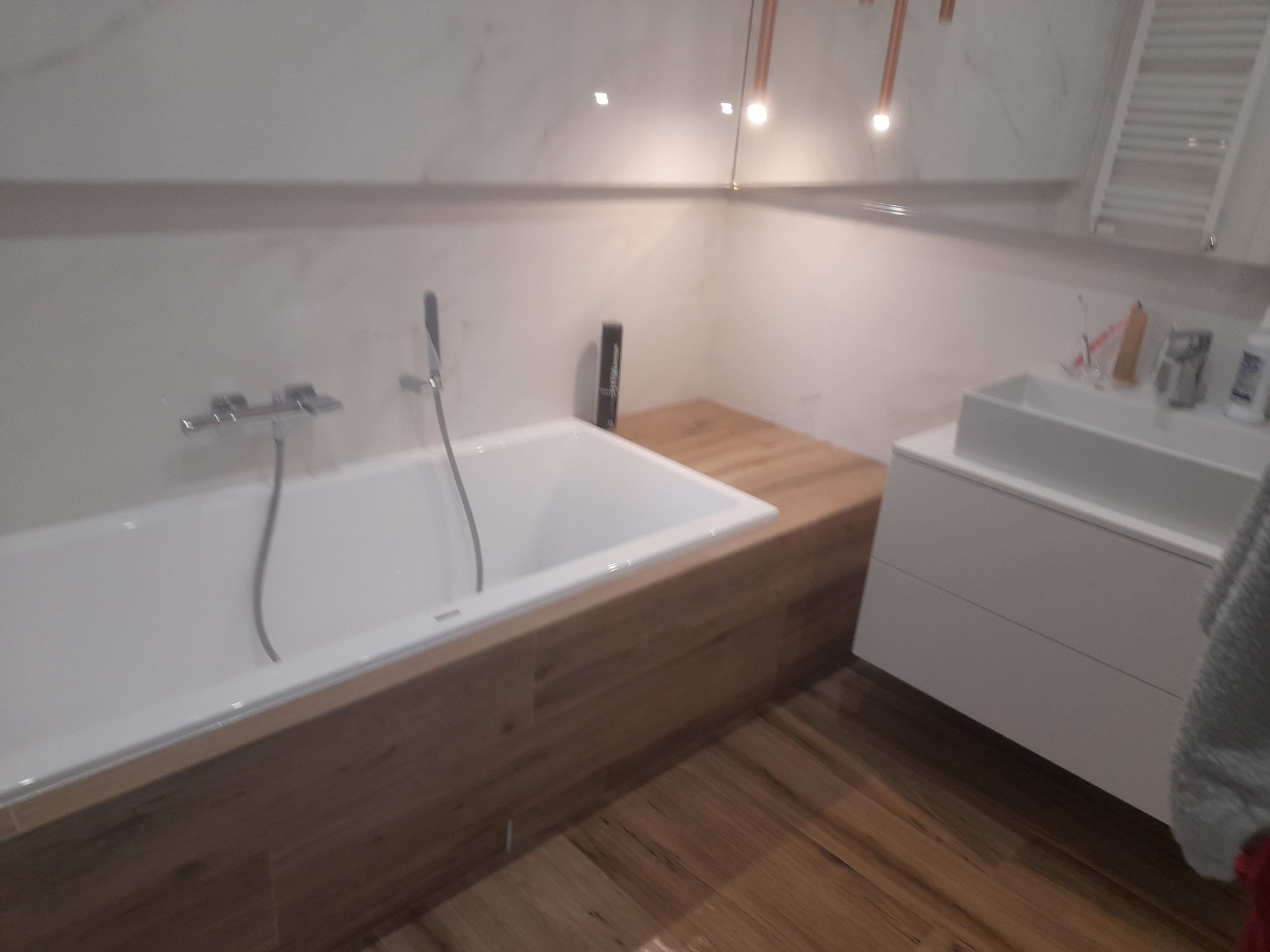 Nowoczesna łazienka z białą wanną obudowaną drewnem, drewnianą podłogą i białą umywalką na szafce. Nad wanną bateria wannowa ze słuchawką prysznicową. Na blacie obok wanny stoi czarna butelka...