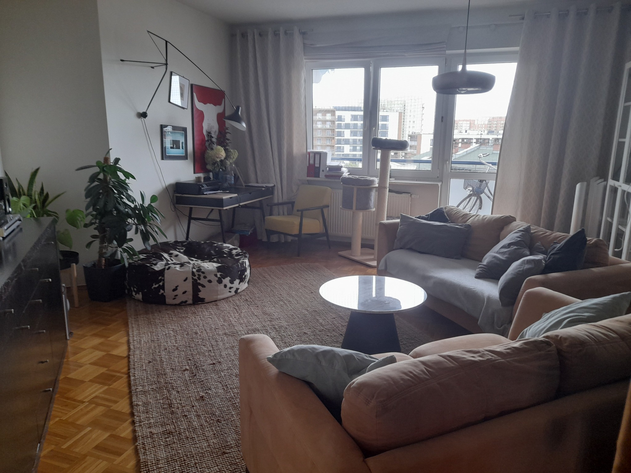 Jasne wnętrze salonu z beżowymi sofami, okrągłym stolikiem kawowym, biurkiem przy oknie i roślinami doniczkowymi, oświetlone naturalnym światłem w dzień.