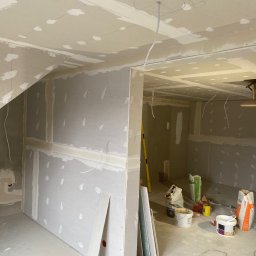 Łukasz Podgórski - Wnętrze w trakcie prac wykończeniowych: ściany i sufit pokryte płytami gipsowo-kartonowymi, widoczne łączenia szpachlowane, wystające przewody elektryczne, worki z zaprawą i narzędzia budowlane...