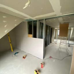 Łukasz Podgórski - Wnętrze w trakcie remontu: montaż ścian działowych z płyt gipsowo-kartonowych na metalowej konstrukcji, widoczna poziomica, wiertarka i kartony po materiałach budowlanych na szarej wylewce.