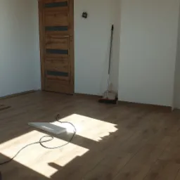 Jasne pomieszczenie z nowo ułożonymi panelami podłogowymi o wzorze drewna, przy ścianie stoi miotła i szufelka, a na podłodze leży biały element z kablem.