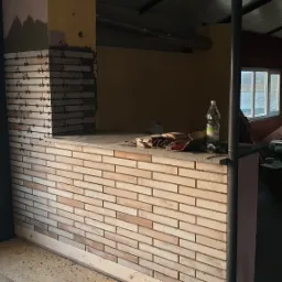 Wnętrze w trakcie remontu z częściowo wykończoną ścianą z cegiełek, widoczne krzyżyki dystansowe, butelka wody mineralnej, narzędzia i rusztowanie.