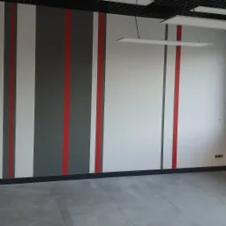 Pomalowane ściany w biurze z pionowymi pasami w kolorach szarym, białym i czerwonym, widoczne lampy sufitowe i okno.