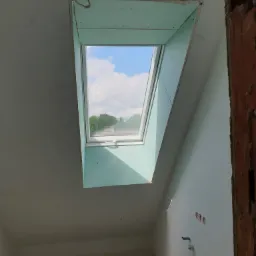 Montaż okna dachowego z obudową z zielonej płyty kartonowo-gipsowej w surowym pomieszczeniu na poddaszu, widoczne instalacje elektryczne i fragment ściany z surowej cegły.