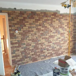 Montaż dekoracyjnej ściany z cegieł w trakcie prac wykończeniowych, widoczne puszki z gruntem, poziomica laserowa, oraz robocze buty na podłodze.