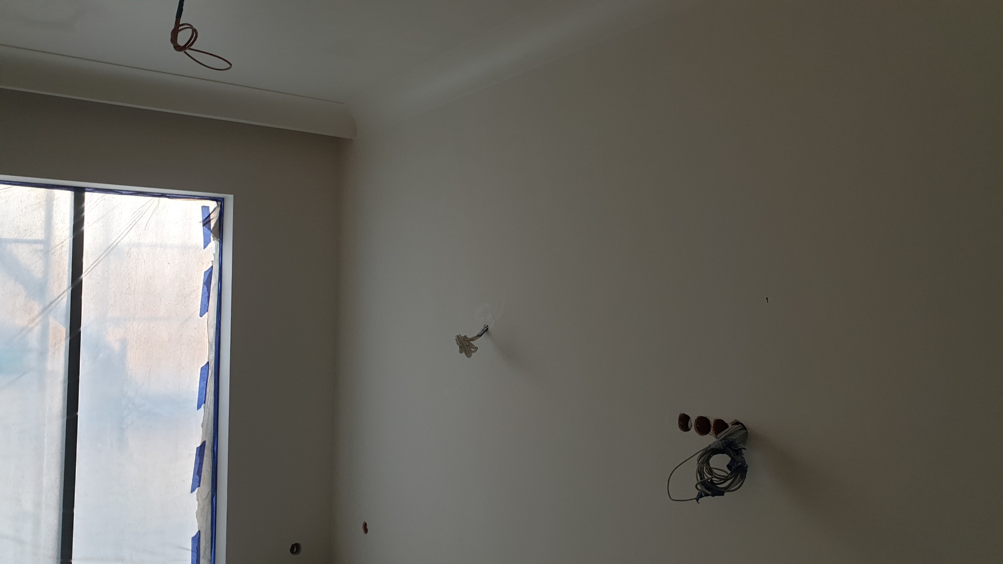 Wnętrze pokoju po remoncie, świeżo pomalowane ściany na jasny odcień, okno zabezpieczone folią malarską, widoczne wystające kable elektryczne.