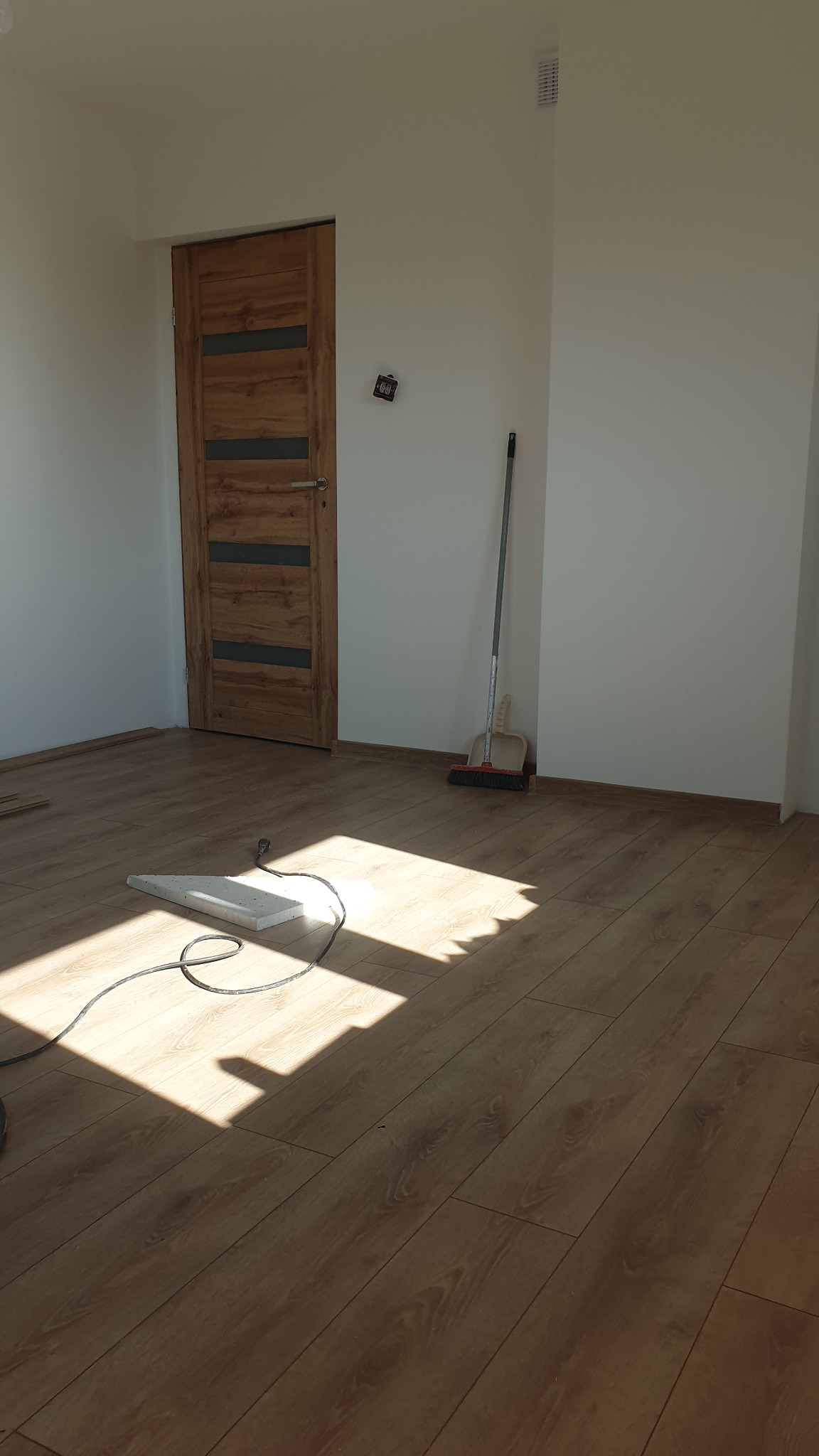 Jasne pomieszczenie z nowo ułożonymi panelami podłogowymi o wzorze drewna, przy ścianie stoi miotła i szufelka, a na podłodze leży biały element z kablem.