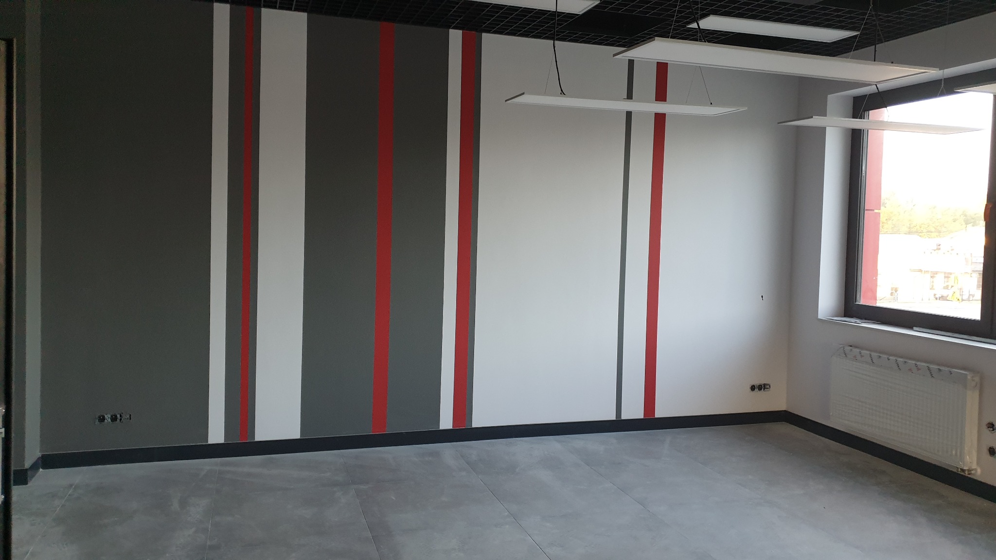 Pomalowane ściany w biurze z pionowymi pasami w kolorach szarym, białym i czerwonym, widoczne lampy sufitowe i okno.