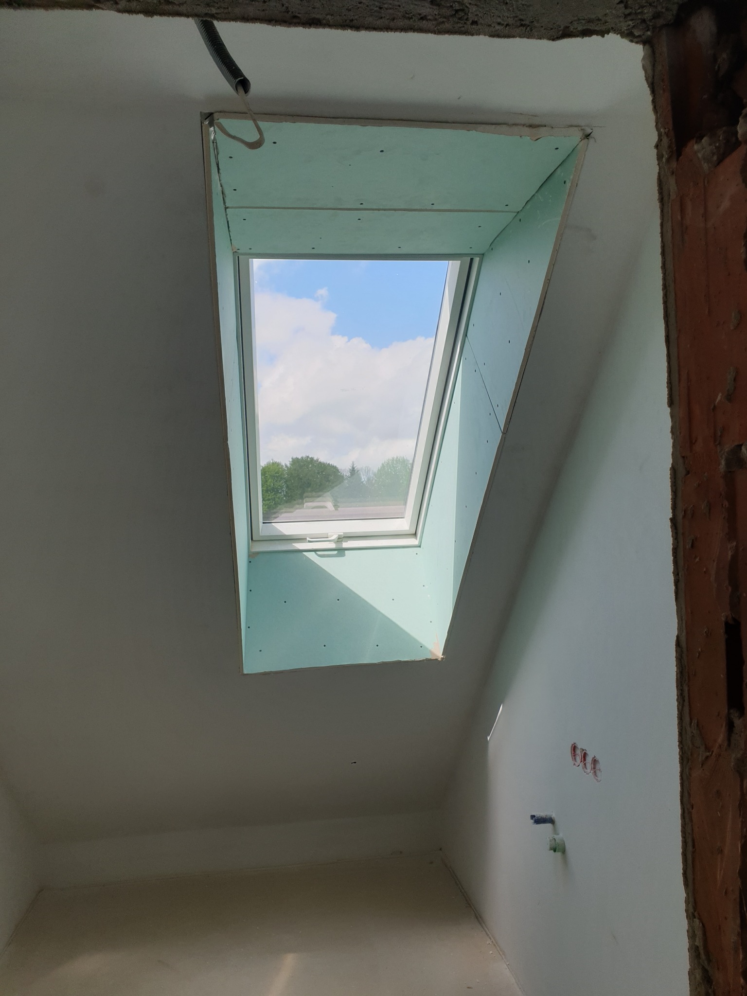 Montaż okna dachowego z obudową z zielonej płyty kartonowo-gipsowej w surowym pomieszczeniu na poddaszu, widoczne instalacje elektryczne i fragment ściany z surowej cegły.